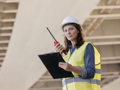 female engineer with a walkie-talkie at the construction site gives instructions. European successful female technologist or fire safety engineer works. foreman in a white helmet and protective yellow jacket on the ladder